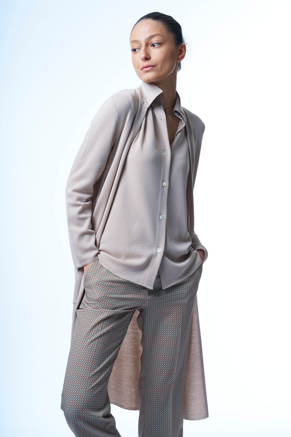 Gilet long beige en 100%mérinos, coupe droite, la maille est resserrée et apporte un beau tombé. Longueur genou. Le gilet manches longues est doté d'un petit col et de 2 poches plaquées. Porté avec une chemise  beige en 100%mérinos.