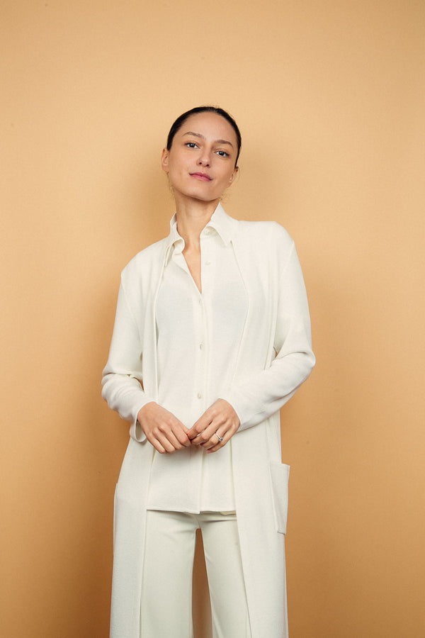 Gilet long blanc en 100%mérinos, coupe droite, la maille est resserrée et apporte un beau tombé. Longueur genou. Le gilet manches longues est doté d'un petit col et de 2 poches plaquées. Porté avec une chemise  blanche en 100%mérinos.