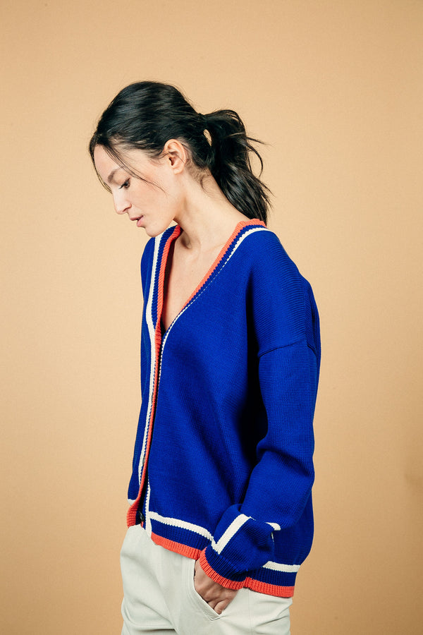 Gilet MALTA bleu Majorelle en coton épais. L'encolure V sur tout son pourtour est rehaussée d'une bande composée de 3 rayures: blanche, bleu, orange également présente sur tout le bas. L'emmanchure est basse et il y a un motif géométrique blanc sur le bas de la manche. 4 boutons.