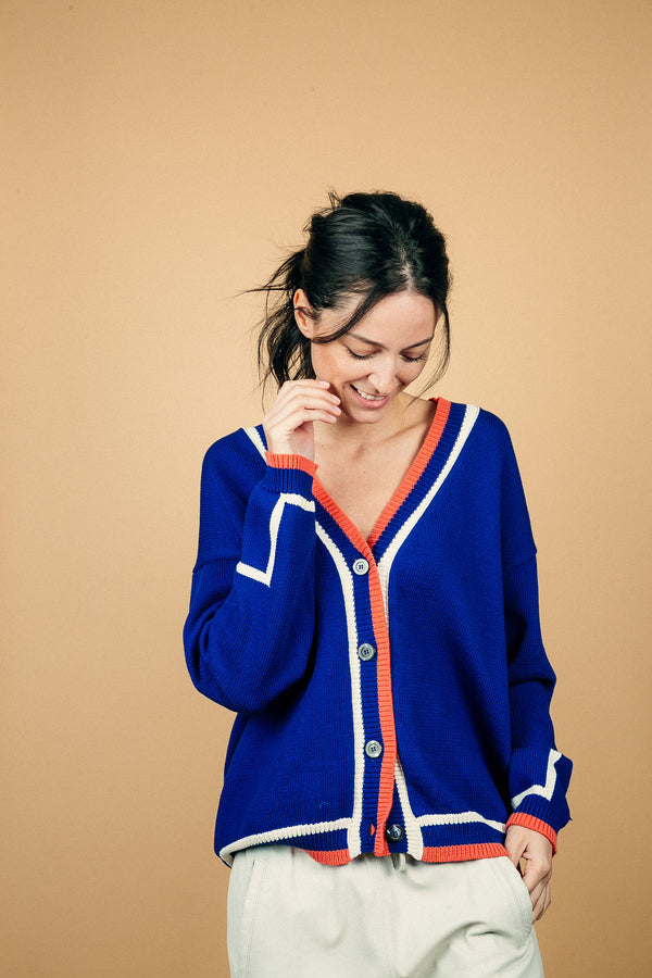 Gilet MALTA bleu Majorelle en coton épais. L'encolure V sur tout son pourtour est rehaussée d'une bande composée de 3 rayures: blanche, bleu, orange également présente sur tout le bas. L'emmanchure est basse et il y a un motif géométrique blanc sur le bas de la manche. 4 boutons.