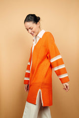 Gilet long orange tricoté au point mousse. Coupe confort, 80% de coton et 15% de cachemire. Les emmanchures sont basses, la manche est large, pas resserrée et a 3 bandes rayées espacées, chacune étant composée d'une ligne écru collée à une ligne taupe. Bande également présente sur tout le pourtour du gilet et sur les fentes de chaque côté. Vue profil.