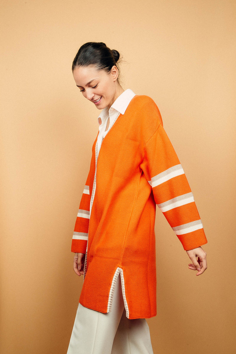 Gilet long orange tricoté au point mousse. Coupe confort, 80% de coton et 15% de cachemire. Les emmanchures sont basses, la manche est large, pas resserrée et a 3 bandes rayées espacées, chacune étant composée d'une ligne écru collée à une ligne taupe. Bande également présente sur tout le pourtour du gilet et sur les fentes de chaque côté. Vue profil.