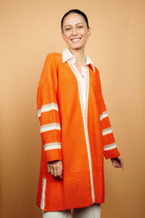 Gilet long orange tricoté au point mousse. Coupe confort, 80% de coton et 15% de cachemire. Les emmanchures sont basses, la manche est large, pas resserrée et a 3 bandes rayées espacées, chacune étant composée d'une ligne écru collée à une ligne taupe. Bande également présente sur tout le pourtour du gilet et sur les fentes de chaque côté.