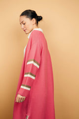 Gilet long rose malabar tricoté au point mousse. Coupe confort, 80% de coton et 15% de cachemire. Les emmanchures sont basses, la manche est large, pas resserrée et a 3 bandes rayées espacées, chacune étant composée d'une ligne écru collée à une ligne taupe. Bande également présente sur tout le pourtour du gilet et sur les fentes de chaque côté. Vue profil.