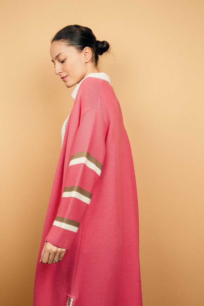 Gilet long rose malabar tricoté au point mousse. Coupe confort, 80% de coton et 15% de cachemire. Les emmanchures sont basses, la manche est large, pas resserrée et a 3 bandes rayées espacées, chacune étant composée d'une ligne écru collée à une ligne taupe. Bande également présente sur tout le pourtour du gilet et sur les fentes de chaque côté. Vue profil.