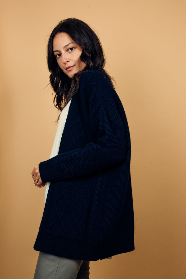 Gilet coloris marine en cachemire, soie et mérinos. Il a 1 grande poche plaquée de chaque côté, la forme est droite, il arrive au niveau des hanches. Il y a un travail de losanges sur le devant et  de point de riz sur les manches et le dos.