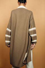 Gilet long taupe tricoté au point mousse. Coupe confort, 80% de coton et 15% de cachemire. Les emmanchures sont basses, la manche est large, pas resserrée et a 3 bandes rayées espacées, chacune étant composée d'une ligne écru collée à une ligne beige. Bande également présente sur tout le pourtour du gilet et sur les fentes de chaque côté. Vue dos.