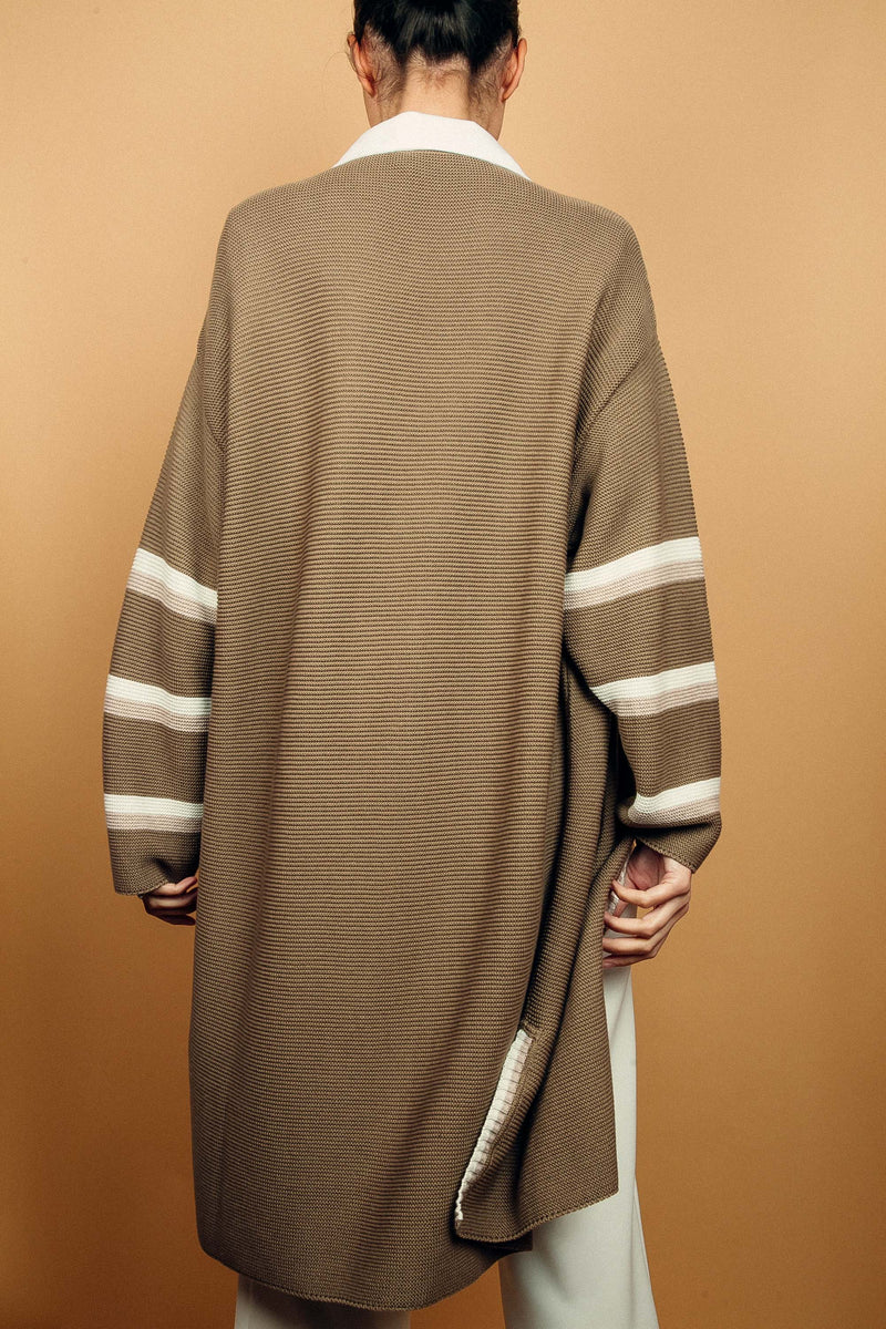 Gilet long taupe tricoté au point mousse. Coupe confort, 80% de coton et 15% de cachemire. Les emmanchures sont basses, la manche est large, pas resserrée et a 3 bandes rayées espacées, chacune étant composée d'une ligne écru collée à une ligne beige. Bande également présente sur tout le pourtour du gilet et sur les fentes de chaque côté. Vue dos.