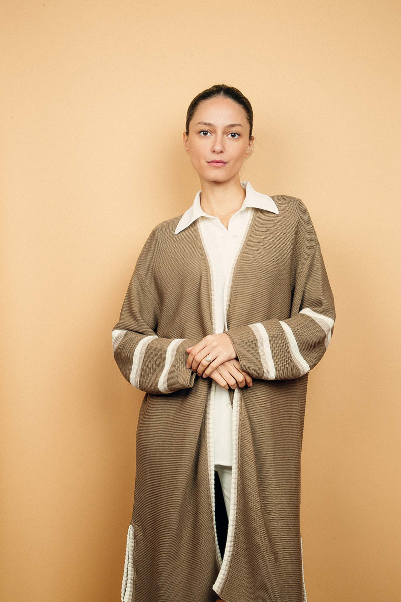 Gilet long taupe tricoté au point mousse. Coupe confort, 80% de coton et 15% de cachemire. Les emmanchures sont basses, la manche est large, pas resserrée et a 3 bandes rayées espacées, chacune étant composée d'une ligne écru collée à une ligne beige. Bande également présente sur tout le pourtour du gilet et sur les fentes de chaque côté.