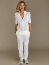 Pantalon pour femme coloris blanc, fluide et léger composé de 94%polyester et de 6% élasthanne. Coupe droite, poches italiennes. Ceinture avec passants, bouton doré.
Vue face.