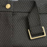 Pantalon de forme bootcut souple et confortable: jambe légèrement appuyée et bas évasé. Le tissu marron est imprimé de petits croisillons beige et vert. La ceinture de ce leggings est recouverte du même tissu, les œillets sont dorés ainsi que le bouton. Esprit années 70's. Le tissu est composé de 69% polyester, 27% rayon, 4% élasthanne. Vue détail de la ceinture.