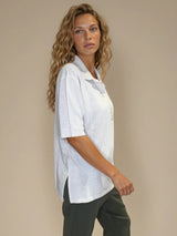 Polo femme d'été coloris beige en 83% viscose et 17% nylon: fluide, coupe large et longue fendue sur les côtés. Le polo a un petit col qui s'ouvre sur 3 boutons, les manches courtes couvrent les avant-bras. Vue profil.