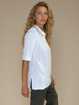 Polo femme d'été coloris blanc en 83% viscose et 17% nylon: fluide, coupe large et longue fendue sur les côtés. Le polo a un petit col qui s'ouvre sur 3 boutons, les manches courtes couvrent les avant-bras. Vue profil.
