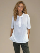 Polo femme d'été coloris blanc en 83% viscose et 17% nylon: fluide, coupe large et longue fendue sur les côtés. Le polo a un petit col qui s'ouvre sur 3 boutons, les manches courtes couvrent les avant-bras. Vue face.