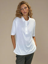 Polo femme d'été coloris blanc en 83% viscose et 17% nylon: fluide, coupe large et longue fendue sur les côtés. Le polo a un petit col qui s'ouvre sur 3 boutons, les manches courtes couvrent les avant-bras. Vue face.