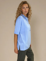 Polo femme d'été coloris ciel en 83% viscose et 17% nylon: fluide, coupe large et longue fendue sur les côtés. Le polo a un petit col qui s'ouvre sur 3 boutons, les manches courtes couvrent les avant-bras. Vue profil.