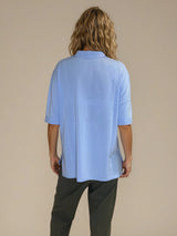 Polo femme d'été coloris ciel en 83% viscose et 17% nylon: fluide, coupe large et longue fendue sur les côtés. Le polo a un petit col qui s'ouvre sur 3 boutons, les manches courtes couvrent les avant-bras. Vue dos.