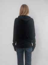 Pull encolure ronde souple avec capuche coloris noir en 100% laine mérinos. Manches longues terminées par un long poignet côtelé avec une ouverture pour y glisser le pouce comme une mitaine. Emmanchures raglan, large bande côtelée sur le bas. Vue dos.