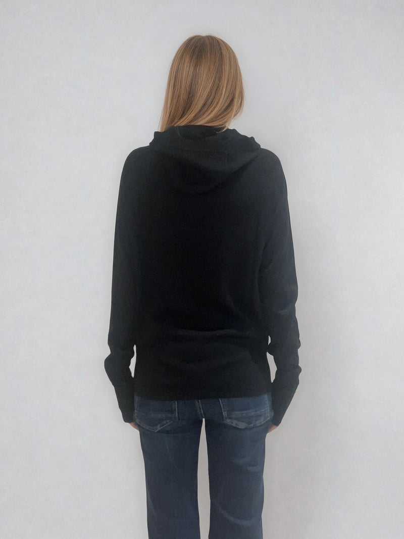 Pull encolure ronde souple avec capuche coloris noir en 100% laine mérinos. Manches longues terminées par un long poignet côtelé avec une ouverture pour y glisser le pouce comme une mitaine. Emmanchures raglan, large bande côtelée sur le bas. Vue dos.