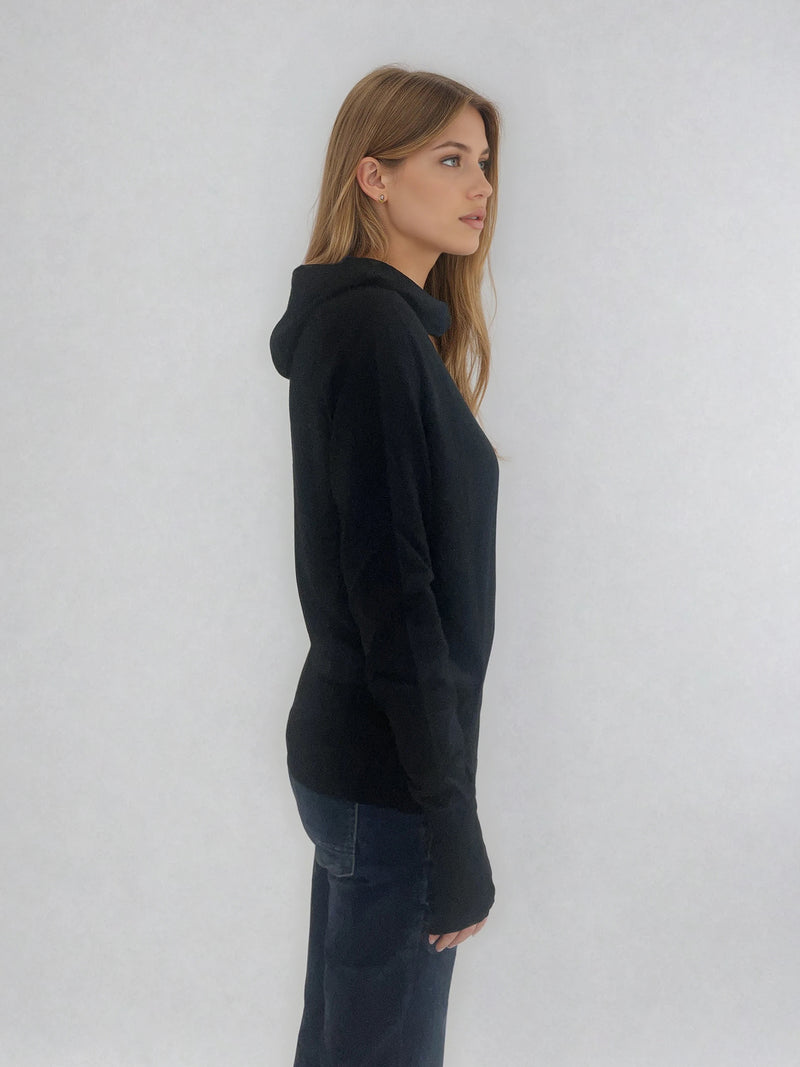 Pull encolure ronde souple avec capuche coloris noir en 100% laine mérinos. Manches longues terminées par un long poignet côtelé avec une ouverture pour y glisser le pouce comme une mitaine. Emmanchures raglan, large bande côtelée sur le bas. Vue profil.