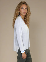 Pull manches longues col rond. Le pull est beige, a un col blanc très arrondi rehaussé d'une bande bleu ciel, détail présent aussi sur le bas des manches. Coupe souple avec une asymétrie sur les côtés.
Composition: 83%viscose et 17% nylon. Vue profil.
