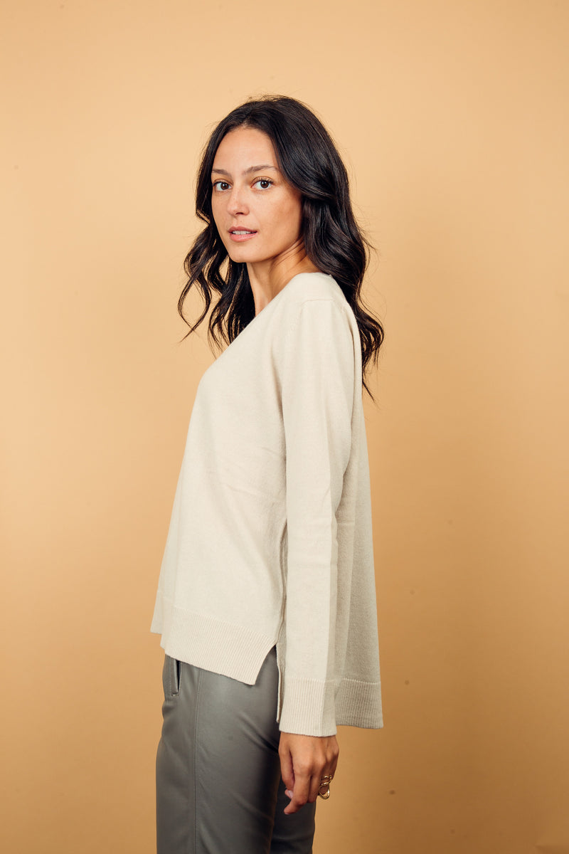 Pull manches longues coloris écru 100% cachemire, col V profond, fendu sur les côtés avec asymétrie: 10 cm dans le dos et 7 cm devant. Finitions bords côtes. 
