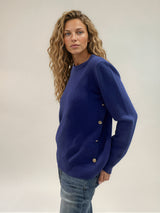 Pull manches longues coloris bleu avec un col rond souple. Le pull a une forme droite. Il y a une rangée de 4 boutons dorés côté droit montée sur une patte du dessous de la manche aux côtes du bas. Composition: 42% cachemire, 8% soie, 50% mérinos.