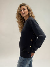 Pull manches longues coloris noir avec un col rond souple. Le pull a une forme droite. Il y a une rangée de 4 boutons dorés côté droit montée sur une patte du dessous de la manche aux côtes du bas. Composition: 42% cachemire, 8% soie, 50% mérinos.