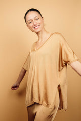 T-shirt long couvrant le bas du dos avec une coupe oversize, col V, manches courtes qui tombent sur les avant-bras, fentes sur les côtés d'environ 15 cm, couture centrale de la pointe du V jusqu'en bas, viscose fluide. Coloris doré miel légèrement brillant.Vue face avec le pantalon en viscose assorti.