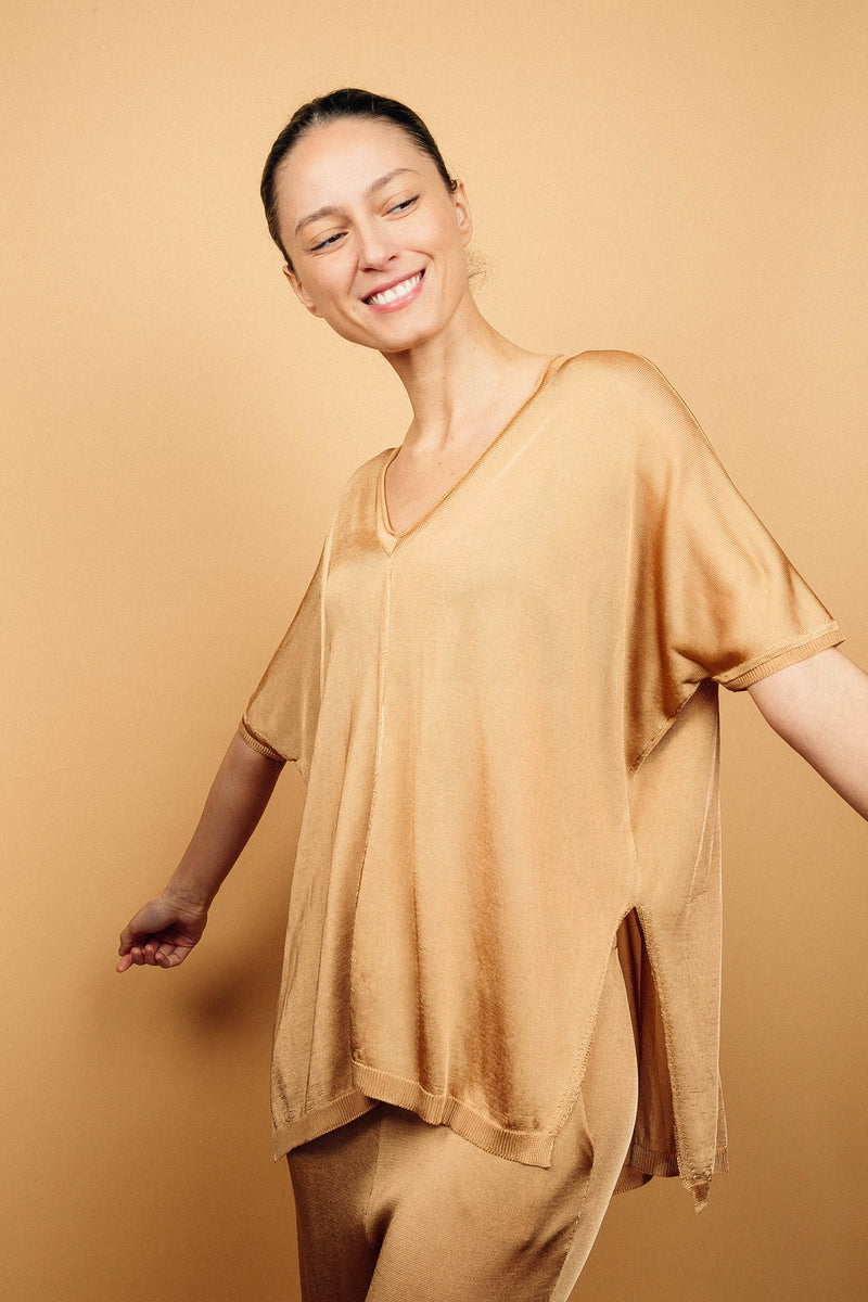 T-shirt long couvrant le bas du dos avec une coupe oversize, col V, manches courtes qui tombent sur les avant-bras, fentes sur les côtés d'environ 15 cm, couture centrale de la pointe du V jusqu'en bas, viscose fluide. Coloris doré miel légèrement brillant.Vue face avec le pantalon en viscose assorti.