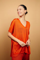 T-shirt long couvrant le bas du dos avec une coupe oversize, col V, manches courtes qui tombent sur les avant-bras, fentes sur les côtés d'environ 15 cm, couture centrale de la pointe du V jusqu'en bas, viscose fluide. Coloris orange uni légèrement brillant. Vue face avec le pantalon en viscose imprimé orange assorti.