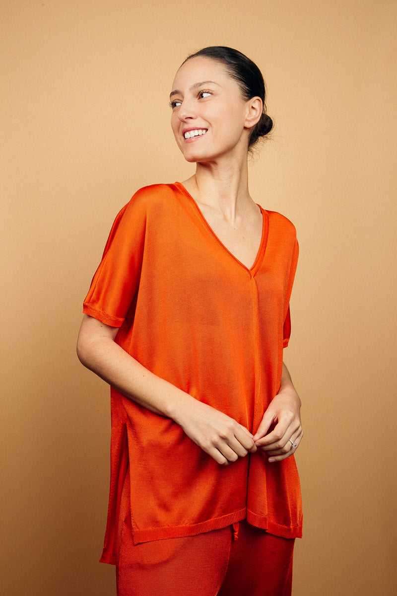 T-shirt long couvrant le bas du dos avec une coupe oversize, col V, manches courtes qui tombent sur les avant-bras, fentes sur les côtés d'environ 15 cm, couture centrale de la pointe du V jusqu'en bas, viscose fluide. Coloris orange uni légèrement brillant. Vue face avec le pantalon en viscose imprimé orange assorti.