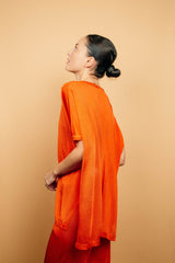 T-shirt long couvrant le bas du dos avec une coupe oversize, col V, manches courtes qui tombent sur les avant-bras, fentes sur les côtés d'environ 15 cm, couture centrale de la pointe du V jusqu'en bas, viscose fluide. Coloris orange uni légèrement brillant. Vue dos avec le pantalon en viscose imprimé orange assorti.