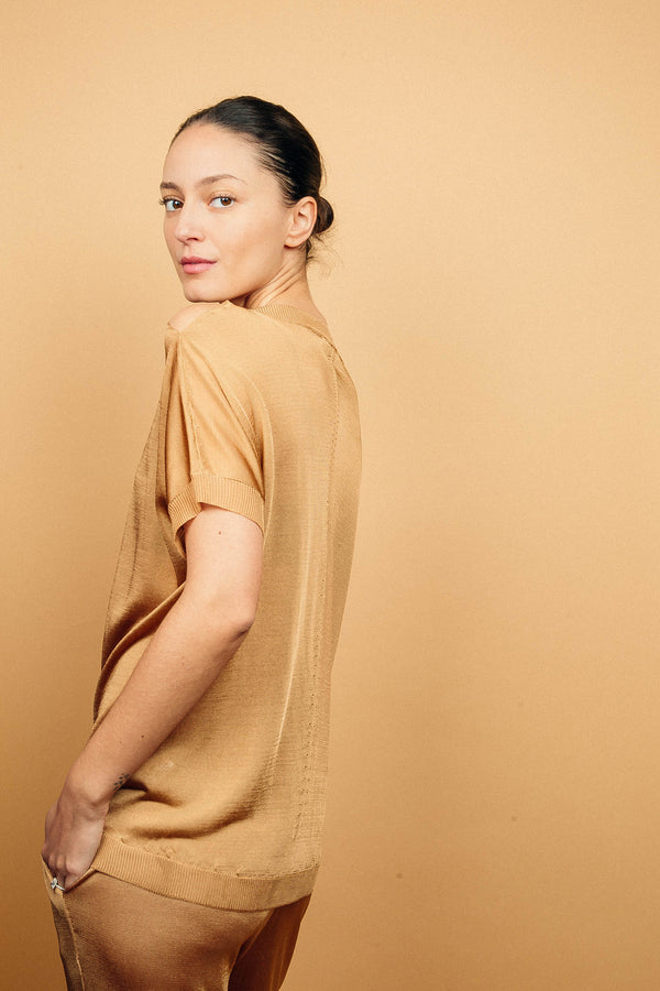 Top fluide en 100% viscose, manches courtes. Il a le haut des épaules dénudé sur 10 cm à partir de l'encolure qui est largement arrondie. La manche recomposée couvre bien l'avant-bras. Coloris doré miel légèrement brillant. Petits points ajourés qui forment une ligne de haut en bas au milieu sur le devant et le dos. Encolure, manches et bas ont des côtes. Peut se porter long ou blousé. Viscose fluide. Il est associé au pantalon en viscose fluide doré miel. Vue profil.