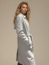 Trench beige 100% polyester avec une coupe droite, resserré par une ceinture. Longueur genoux. Bavolet et fente dans le dos. Fermeture avec zip et patte de boutonnage pressions. Vue profil.