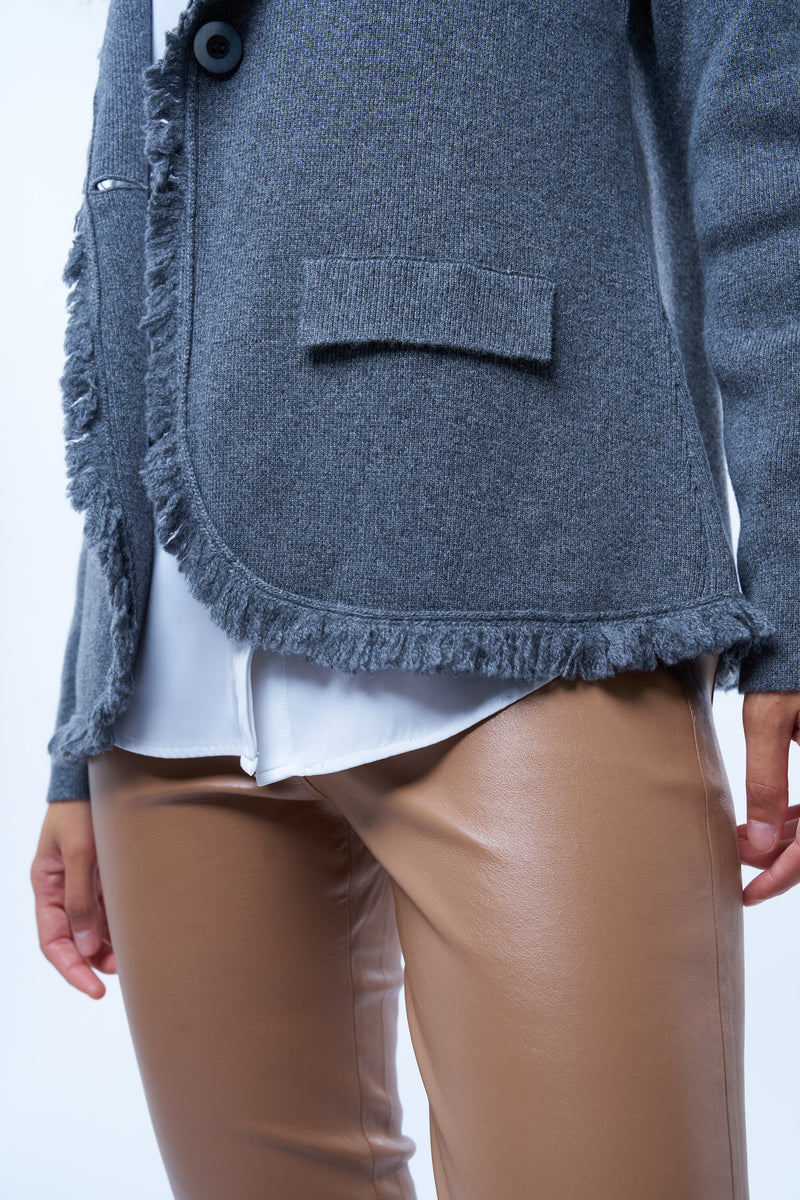 Veste manches longues grise en cachemire, laine et mérinos. La veste est bordée tout le long du col tailleur et en bas de petites franges grises en laine, se ferme avec 1 bouton et a  2 fausses poches sur les côtés. Coupe droite. Vue détail franges du bas.