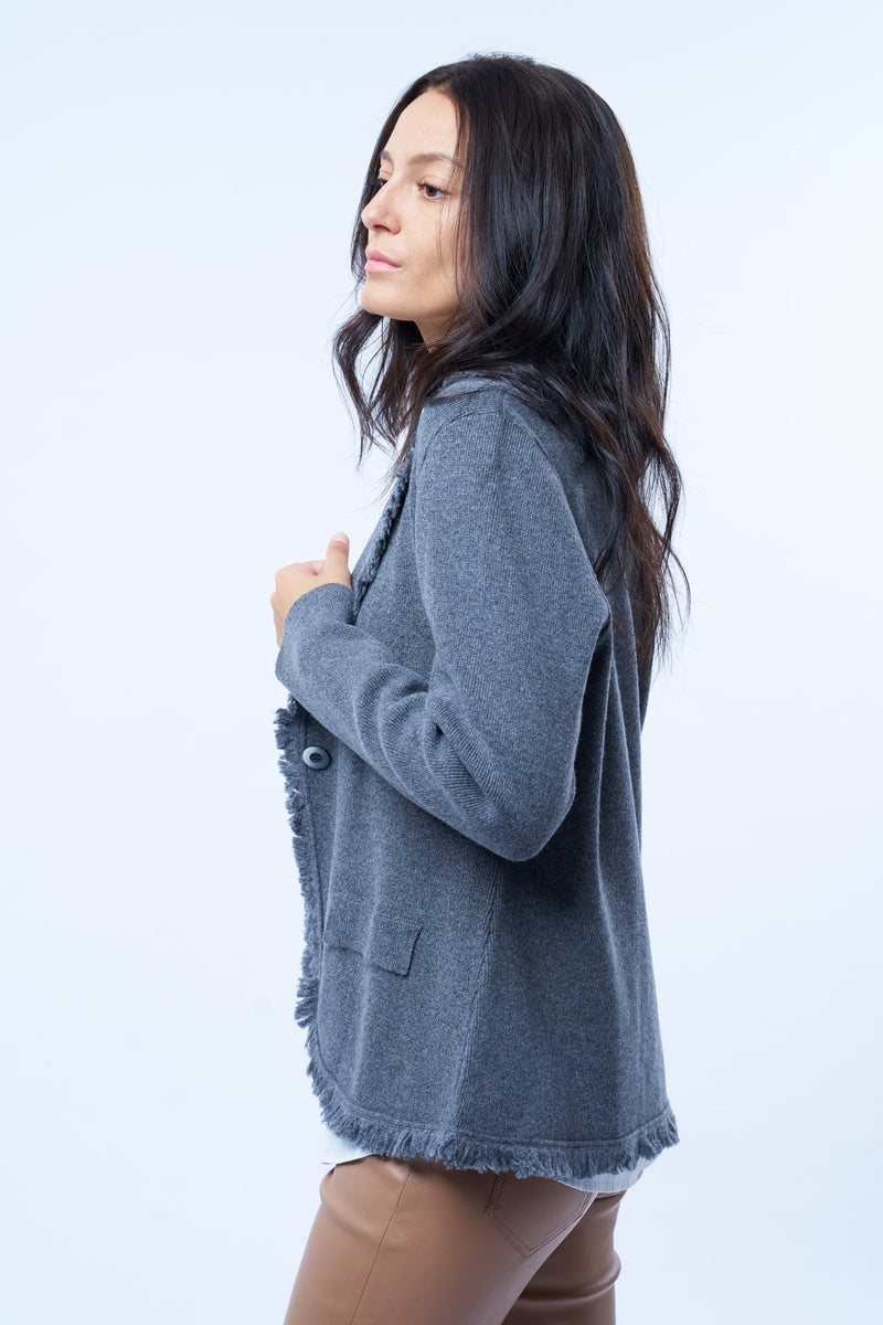 Veste manches longues grise en cachemire, laine et mérinos. La veste est bordée tout le long du col tailleur et en bas de petites franges grises en laine, se ferme avec 1 bouton et a  2 fausses poches sur les côtés. Coupe droite. Vue profil.