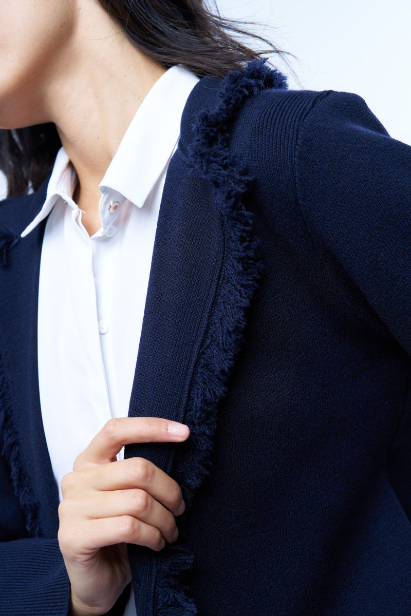 Veste manches longues marine en cachemire, laine et mérinos. La veste est bordée tout le long du col tailleur et en bas de petites franges marines en laine, se ferme avec 1 bouton et a  2 fausses poches sur les côtés. Coupe droite. Vue détail franges sur le col tailleur.